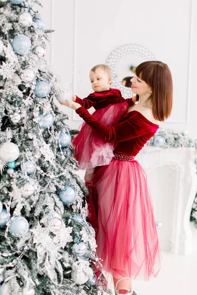 Felice giovane madre dai capelli rossi e piccola figlia, indossa eleganti abiti rossi vino, divertirsi e decorare l'albero di Natale in camera luce accogliente — Foto Stock