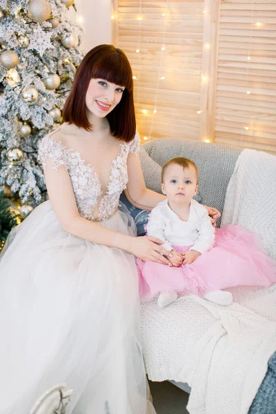 Красива мати зі своєю дитиною, одягнена в модні елегантні сукні, сидить на стильному сірому дивані на фоні ялинки, вогні та новорічні прикраси — стокове фото