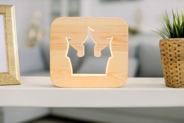 Dekoratif, ahşap gece lambası, şato kulesi resmi, çiçek saksısı ve fotoğraf çerçeveli ışıklı masa. Arka planda gri kanepe olan rahat bir ev.