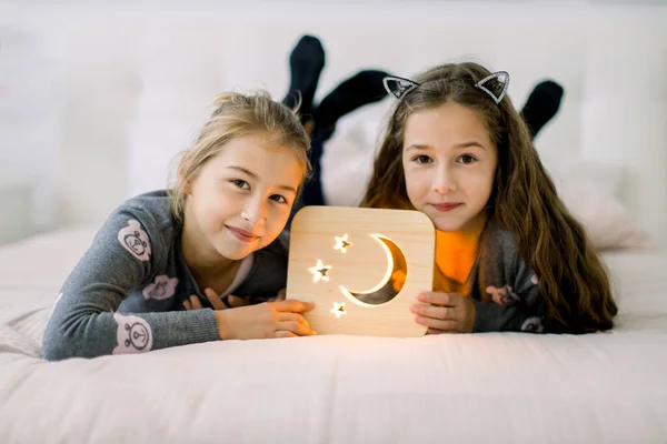 Twee mooie kleine meisjes zusjes, die thuis op bed liggen en genieten van hun tijd spelend met houten nachtlampje met maan- en sterrenbeeld. Prettige tijd thuis. Gezellige huisdecoraties — Stockfoto