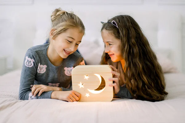 Twee mooie kleine meisjes zusjes, die thuis op bed liggen en genieten van hun tijd spelend met houten nachtlampje met maan- en sterrenbeeld. Prettige tijd thuis. Gezellige huisdecoraties — Stockfoto