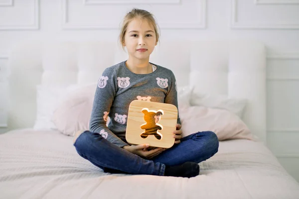 Indoor portret van mooie school meisje in casual huishoudelijke kleding, zittend op het bed in lotus positie en met stijlvolle houten nachtlamp met beer foto. — Stockfoto