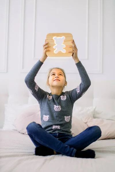 Indoor portret van mooie school meisje in casual huishoudelijke kleding, zittend op het bed in lotus positie en met stijlvolle houten nachtlamp met beer foto onder haar hoofd. — Stockfoto