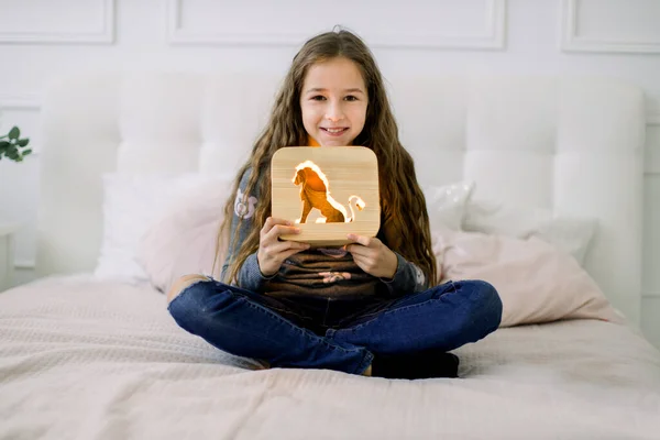 Indoor portret van mooie school meisje in casual huishoudelijke kleding, zittend op het bed in lotus positie en met stijlvolle houten nachtlamp met beer foto. — Stockfoto