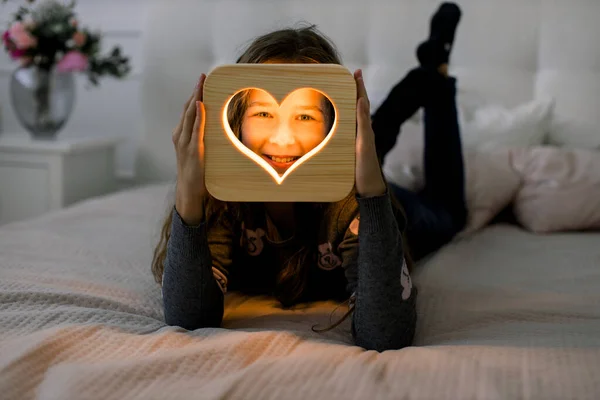 Klein schattig meisje, liggend op het bed in gezellige slaapkamer, en het maken van verschillende emoties, terwijl het houden van houten nachtlamp met hart uitgesneden beeld. Houten interieur — Stockfoto