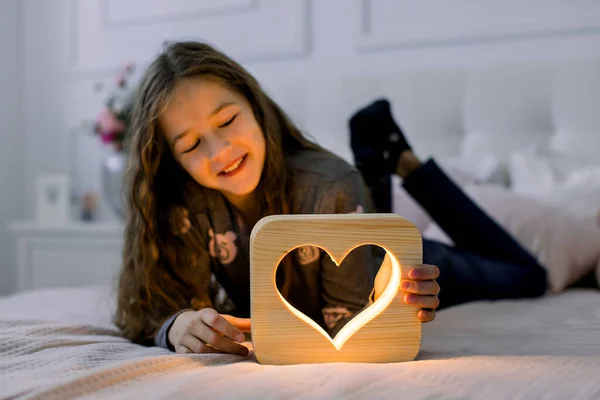 Çocuklar, duygular ve gece lambaları konsepti. Güzel bir kız öğrencinin yatay çekimleri, yatağında yatarken ve eliyle ahşap bir gece lambasıyla eğlenirken. — Stok fotoğraf