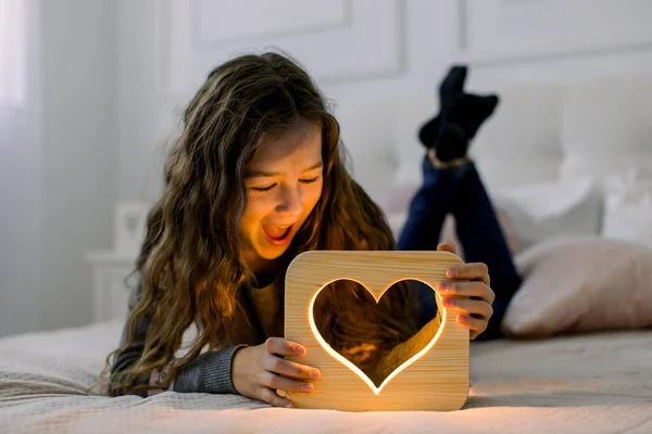 Kinderen, emoties en nachtlampenconcept. Indoor horizontale schot van mooie kind schoolmeisje, liggend op het bed thuis en plezier hebben met stijlvolle hand gemaakte houten nachtlamp met hart foto — Stockfoto