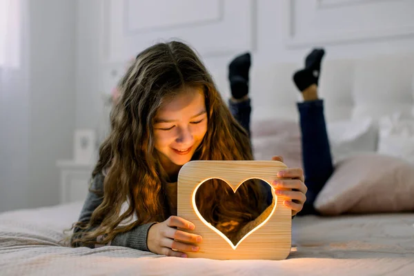 Çocuklar, duygular ve gece lambaları konsepti. Güzel bir kız öğrencinin yatay çekimleri, yatağında yatarken ve eliyle ahşap bir gece lambasıyla eğlenirken. — Stok fotoğraf