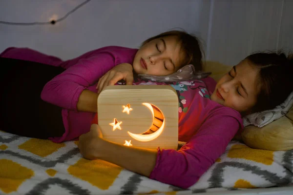 Slaap lekker, goede nacht en nacht lampen promo concept. Close-up van twee prachtige 10-jarige meisjes zussen, samen liggend in het gezellige bed, met houten nachtlampje, en slapen of slapen — Stockfoto