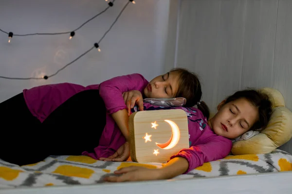 Tatlı rüyalar, iyi geceler ve gece lambaları tanıtım konsepti. İki güzel 10 yaşındaki kız kardeşin, rahat bir yatakta, ahşap bir gece lambasıyla uzanması, uyuması ya da uyuması. — Stok fotoğraf