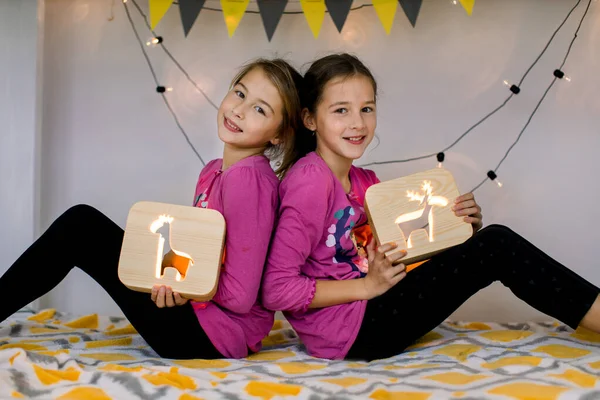 Mutlu iki sevimli kız kardeş 10 yaşında, çocuk odasında süslü bir ranzada, sırt sırta oturuyorlar ve kesilmiş resimli ahşap gece lambaları tutuyorlar.. — Stok fotoğraf