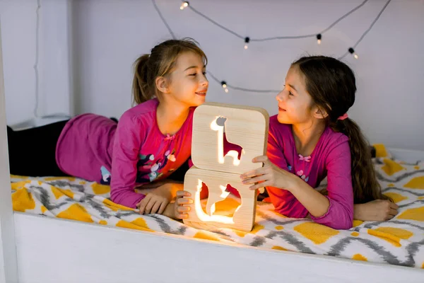 Twee schattige blanke kinderen, mooie kleine meisjes zusjes, het dragen van roze shirts, liggend op een helder kinderbed stapelbed en spelen met stijlvolle houten nachtlampjes met uitgesneden foto 's — Stockfoto