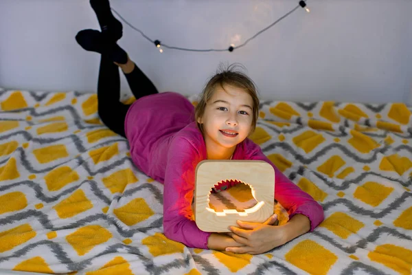 Close-up horizontaal schot van gelukkig glimlachen 10 jaar oud meisje, liggend op gezellig bed, met mooie houten nachtlamp met egel foto — Stockfoto