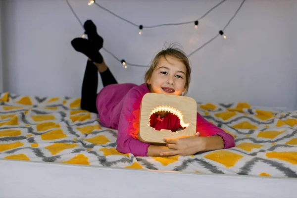 Yatay çekimde mutlu gülümseyen 10 yaşındaki kız, rahat bir yatakta uzanıyor, elinde kirpi resimli güzel bir ahşap gece lambası tutuyor. — Stok fotoğraf