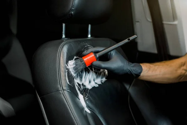 Close up cropped image of washing car interior and seat by a soft brush with foam. Young male worker in black protective gloves cleaning car seat with foam and brush