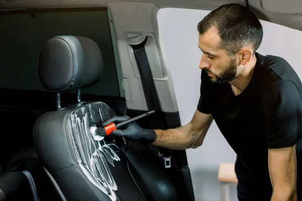 Feche o retrato horizontal do jovem belo trabalhador masculino em t-shirt preta e luvas, limpando o assento dianteiro do carro com espuma especial e escova macia. Detalhes do carro, desinfecção, limpeza — Fotografia de Stock