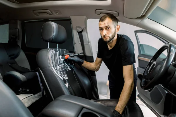 Concepto de detalle y limpieza de coches. Joven, trabajador de lavado de coches, con guantes de goma protectora, la limpieza de la silla de coche del vehículo moderno, utilizando cepillo especial y espuma, mirando a la cámara —  Fotos de Stock