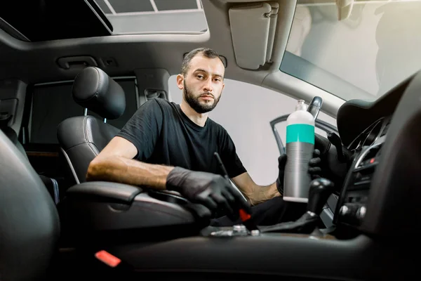 Jovem concentrado trabalhador de serviço de carro masculino limpeza de poeira do interior do carro de luxo, usando escova especial e produto profissional de cuidados de plástico em garrafa. Detalhes do carro, cuidados interiores. Foco no rosto — Fotografia de Stock