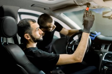 Profesyonel araba temizleme servisi. İki beyaz adamın yan görüntüsü, modern arabanın içini temizliyor, direksiyon simidi ve mikrofiber bez ve özel fırçayla gösterge paneli