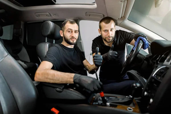 Retrato de dois homens bonitos positivos, serviço de automóvel e trabalhadores detalhando, vestindo roupas e luvas pretas, mostrando polegares até a câmera, enquanto a limpeza interior do veículo moderno — Fotografia de Stock