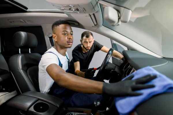 Carro profissional de limpeza e detalhamento conceito. Dois trabalhadores masculinos multirraciais de uniformes, lavando o interior do carro com escova, microfibra e detergente na estação de serviço. Foco no rosto do homem africano — Fotografia de Stock