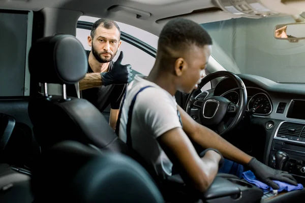 Dois trabalhadores do sexo masculino multiétnico limpeza interior do veículo com pano especial e escova, no serviço de carro detalhando estação. Concentre-se no homem caucasiano atrás, mostrando seu polegar para a câmera — Fotografia de Stock