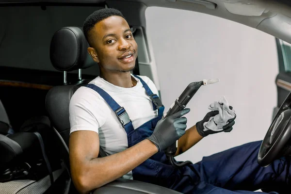 Car steam cleaning, disinfection. Handsome young African American guy auto service worker, wearing white t-shirt and overalls, sitting inside of modern car and posing with steam cleaner and cloth