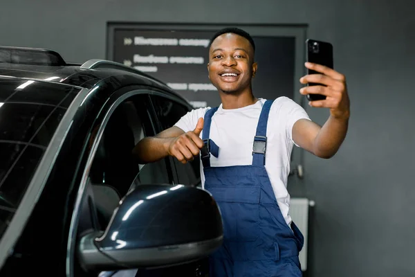Tampan tersenyum tersenyum Afrika laki-laki pekerja auto service, mengenakan t-shirt putih dan biru overall, berdiri di dekat mobil hitam modern, membuat foto selfie di smartphone, menunjukkan jempol up — Stok Foto