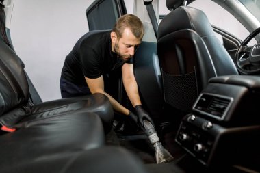 Car wash and detailing service. Male worker in black uniform and gloves, making chemical cleaning process with a vacuum cleaner and foam, releasing car floor from dust and sand
