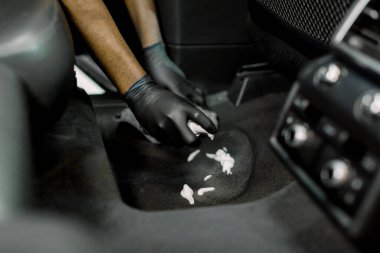 Cropped close up of hands of professional male detailer in black gloves, cleaning the carpet of car interior, applying disinfecting cleaning foam