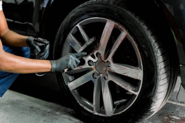 Oto yıkama servisi. Siyah koruyucu eldivenli erkek ellerinin yakın plan görüntüsü, lüks arabanın alaşımlı tekerlek jantlarını temizleme atölyesinde dökme tekerlekler için özel bir fırça ile birlikte