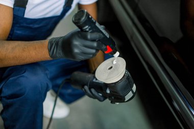 Cropped image of African man, auto service worker, wearing white t-shirt and blue overalls, putting special polish wax or cream on the orbital polisher to polish the black car behind clipart