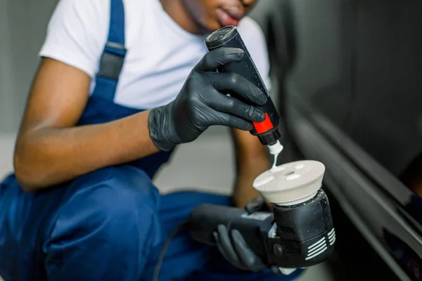 将非洲男性汽车维修人员的手像剪得很近，在轨道抛光机上倒入特殊的抛光蜡或奶油，在背景上擦亮黑色汽车 — 图库照片