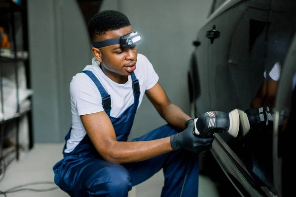 Auto detailing service, polishing of the car. Side view of young African American man worker in t-shirt and overalls, polishing black car body and door with orbital polisher and wax