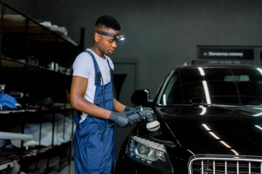 Araba temizliği ve cilalama konsepti. Profesyonel siyah erkek araba servisi çalışanı, el yörüngesi cilalayıcısı, ve oto tamirhanesinde siyah lüks araba cilalıyor.