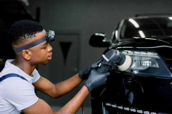 Detallado y pulido del faro del coche. Joven trabajador de piel oscura en guantes uniformes y protectores, con pulidor orbital en auto servicio de pulido de luces de coche — Foto de Stock