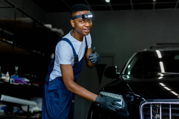 Alegre trabajador africano, con guantes uniformados y protectores, limpiando y frotando el exterior de un automóvil usando esponja. Trabajador profesional puliendo un coche con cera de coche, concepto de cuidado automático. — Foto de Stock