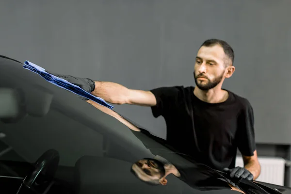 Πλύσιμο και καθαρισμός αυτοκινήτων στο επαγγελματικό πρατήριο αυτοκινήτων. Βολή του όμορφος γενειοφόρος νεαρός εργάτης σε μαύρο t-shirt και γάντια, τον καθαρισμό του αυτοκινήτου παρμπρίζ με μπλε πανί μικροϊνών — Φωτογραφία Αρχείου