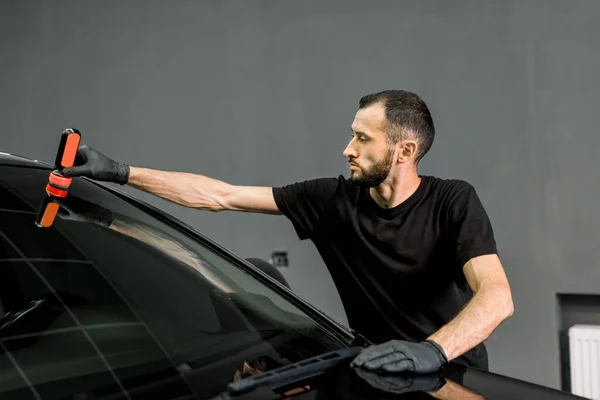 Hombre trabajador en uniforme negro y guantes de protección, en un servicio profesional de lavado de coches, aplicando recubrimiento contra la lluvia en un parabrisas de lujo coche negro — Foto de Stock