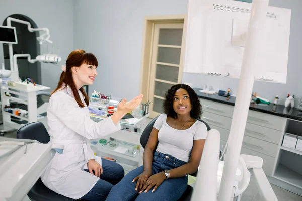 Giovane paziente donna africana seduta sulla sedia di odontoiatria e guardando sullo schermo digitale con l'immagine dei suoi denti. Una dottoressa caucasica che spiega i modi di trattamento. Cure dentali — Foto Stock