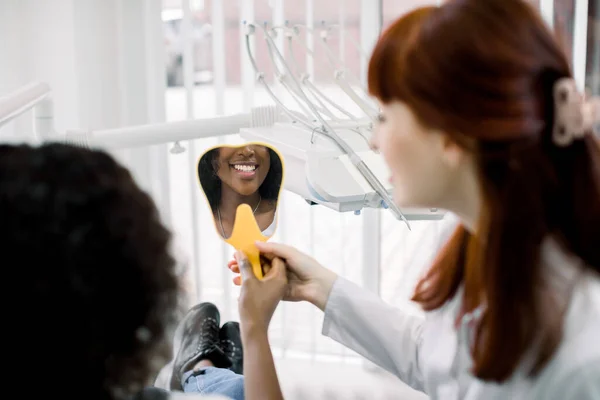 Visão traseira da bela mulher africana paciente e branca dentista feminina, verificando o sorriso após a cura ou clareamento dos dentes na clínica odontológica. Foco no espelho com reflexão de sorriso — Fotografia de Stock