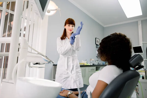 Odontología, cuidado bucal, revisión dental. Mujer joven caucásica dentista, con abrigo blanco, con guantes médicos azules y preparándose para el examen de los dientes de su paciente africana —  Fotos de Stock