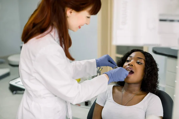 У стоматологічному кабінеті молода африканка-пацієнтка, яка відчиняє рот, посміхаючись жінці-стоматологу в уніфікованих і синіх латексних рукавичках, перевіряє стан зубів, використовуючи зубне дзеркало. Концепція очного догляду — стокове фото
