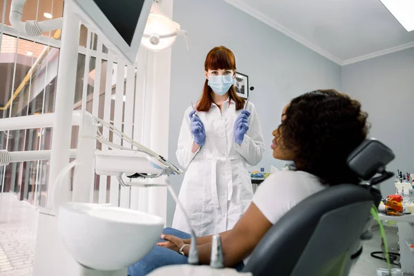 Concepto dental. Odontóloga bastante femenina con abrigo blanco, máscara protectora y guantes de látex azul, muestra instrumentos dentales a la cámara y a su joven paciente africana sentada en la silla dental — Foto de Stock