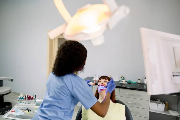 Female African American dentist doing tooth filling procedure with dental curing UV light in clinic. European girl patient in the dental clinic. Teeth whitening UV lamp with photopolymer composition.