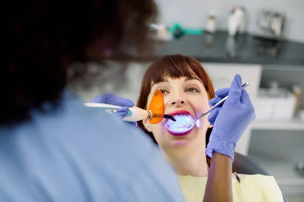 Крупный план лица молодой женщины, проходящей стоматологическое лечение. Обратный вид африканской женщины-стоматолога, делающей стоматологическое лечение с помощью отверждения ультрафиолетовой лампы. Реставрация зубов с заполнением и полимеризацией лампы — стоковое фото