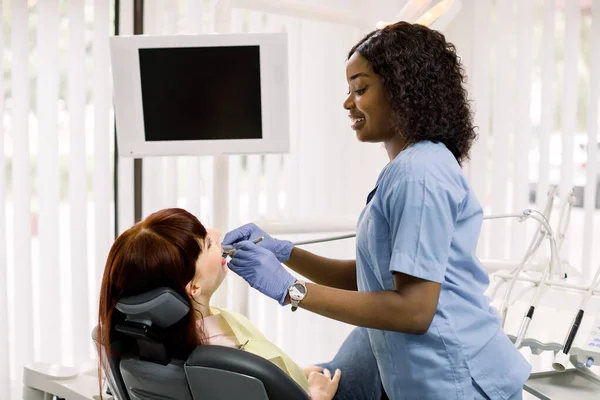 Pretty African female dentist wearing gloves, performing dental check up and treatment on patients teeth. Young woman lying in chair, receiving dental treatment with mouth open