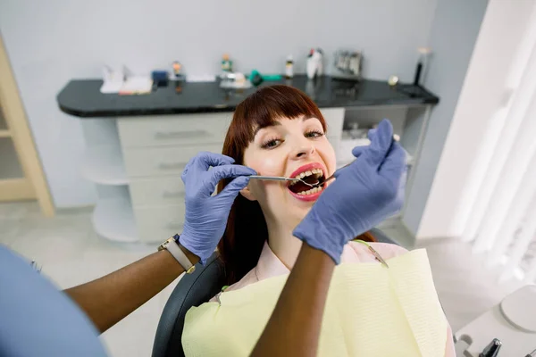 Közelkép felső látószögből egy vonzó európai nő arcát, akit a fogorvos ellenőriz a klinikán. Női afrikai fogorvos, aki fogorvosi eszközökkel vizsgál meg egy beteget.. — Stock Fotó