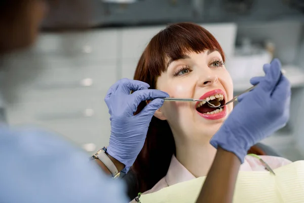 Conceito odontológico e de saúde. Uma foto de perto de uma paciente bonita a fazer um exame dentário na clínica dentária. Mãos de dentista americano africano feminino em luvas azuis examinando dentes de paciente — Fotografia de Stock
