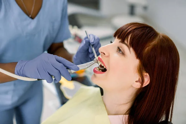 Close-up vista lateral rosto retrato de muito jovem mulher caucasiana paciente tendo tratamento dentário em consultório odontológico. Mãos de dentista africano em luvas segurando ferramentas dentárias — Fotografia de Stock